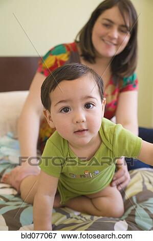 Stock Photo - Hispanic mother watching son crawl on bed. Fotosearch