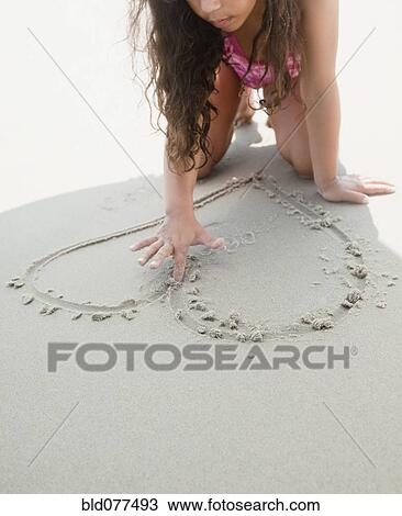 Stock Image - Hispanic girl drawing heart shape in sand. Fotosearch