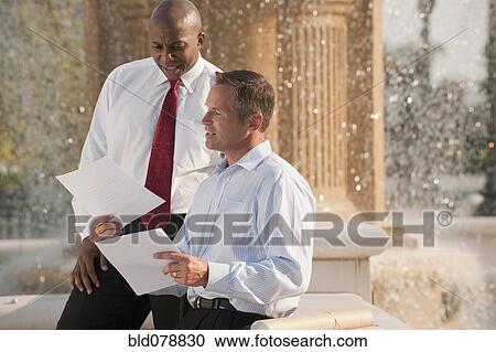 Stock Image - Businessmen reviewing paperwork near fountain. Fotosearch