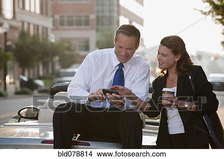 Picture - Caucasian businessman sitting on car text messaging with co-worker. Fotosearch