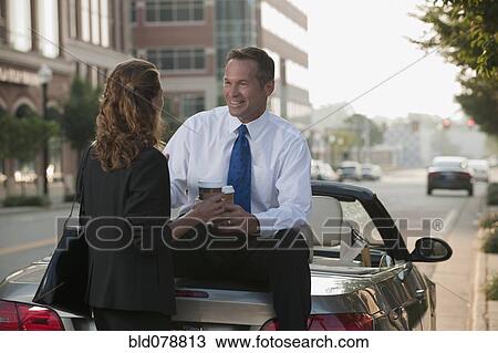 Stock Image - Caucasian businessman sitting on car talking to co-worker. Fotosearch