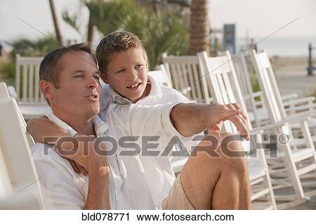 Stock Image - Caucasian father and son sitting in rocking chairs on beach. Fotosearch