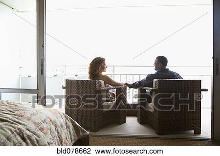 Stock Image - Couple sitting in armchairs and holding hands on urban balcony. Fotosearch