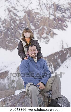 Stock Photograph - Hispanic father and daughter sitting on rock in snow. Fotosearch