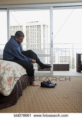 Stock Image - Mixed race man putting socks and shoes on at end of bed. Fotosearch