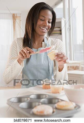 Stock Image - Mixed race woman spreading frosting onto cupcakes. Fotosearch