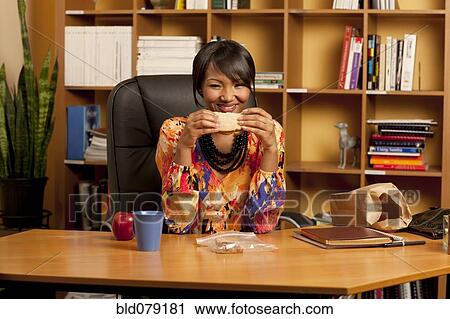 Stock Image - Mixed race businesswoman eating lunch at desk. Fotosearch