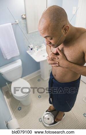 Stock Image - Mixed race man weighing himself on bathroom scale. Fotosearch