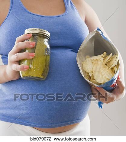 Stock Image - Pregnant Hispanic woman holding pickles and potato chips. Fotosearch