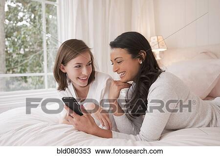 Stock Image - Friends laying on bed looking at cell phone. Fotosearch