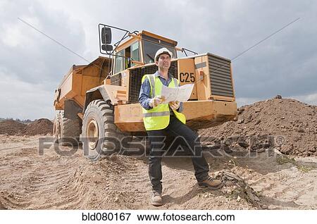 Stock Photo - Hispanic construction worker with blueprints near dump truck. Fotosearch