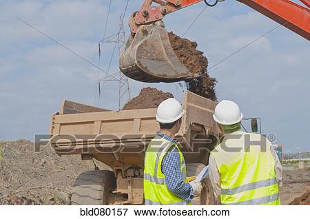 Stock Photo - Hispanic construction workers watching dirt falling into dump truck. Fotosearch