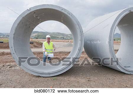 Stock Image - Hispanic worker standing near large cement pipes. Fotosearch