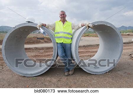 Stock Image - Hispanic worker standing near large cement pipes. Fotosearch