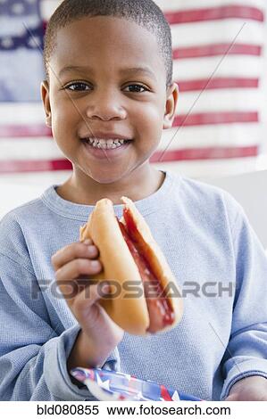 Stock Photography - African American boy eating hot dog in front of American flag. Fotosearch