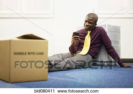 Stock Image - Black businessman sitting on floor text messaging. Fotosearch