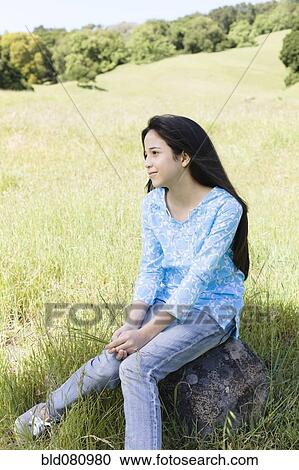 Stock Image - Hispanic girl sitting on rock in field. Fotosearch