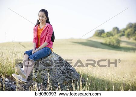 Stock Image - Hispanic girl sitting on rock in field. Fotosearch