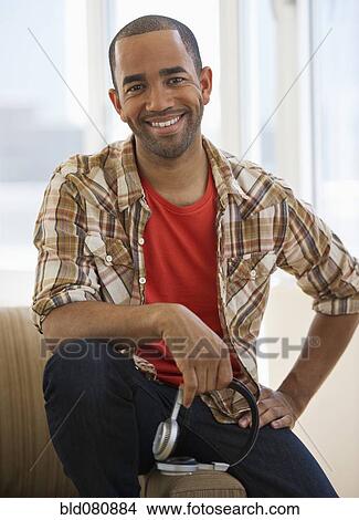 Mixed race man sitting on sofa holding headphones View Large Photo Image Picture - Mixed race man sitting on sofa holding headphones. Fotosearch