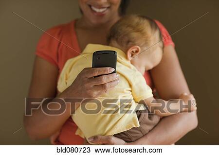 Stock Image - Mother text messaging while holding sleeping child. Fotosearch