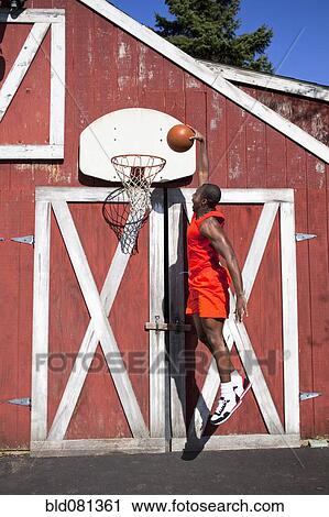 Stock Image - Black man dunking basketball outside barn. Fotosearch