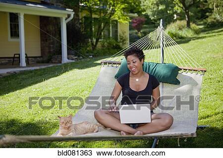 Stock Image - Black woman sitting in hammock with laptop and cat. Fotosearch