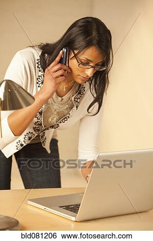 Stock Photograph - Mixed race woman talking on cell phone and using laptop. Fotosearch
