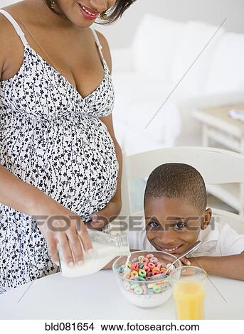 Picture - Black boy smiling as pregnant mother pours milk on cereal. Fotosearch
