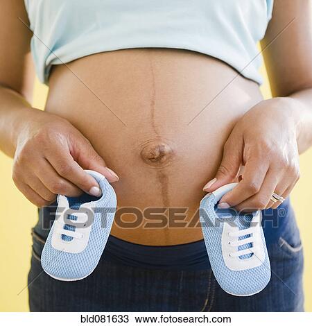 Stock Image - Close up of pregnant Black woman holding blue baby booties to stomach. Fotosearch