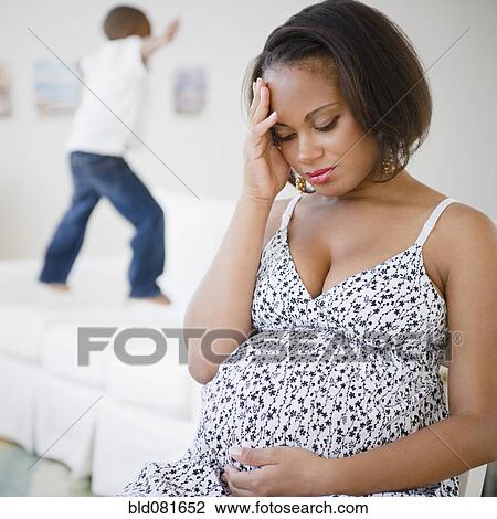 Stock Image - Pregnant Black mother sitting with head in hands while son jumps on bed. Fotosearch