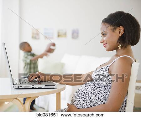 Stock Image - Pregnant Black mother using laptop while son plays with football. Fotosearch