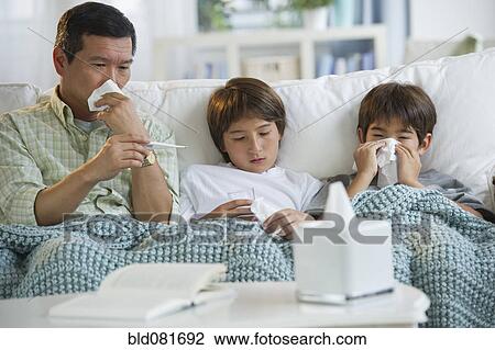 Stock Image - Sick father and sons blowing noses and checking temperature on living room sofa. Fotosearch