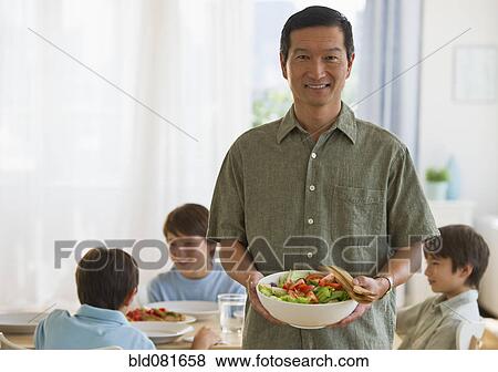 Stock Photo - Smiling father holding salad with sons at table in background. Fotosearch