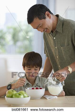 Stock Image - Smiling father pouring milk on son's cereal. Fotosearch