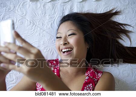 Stock Image - Smiling mixed race teenage girl laying on bed and text messaging on cell phone. Fotosearch
