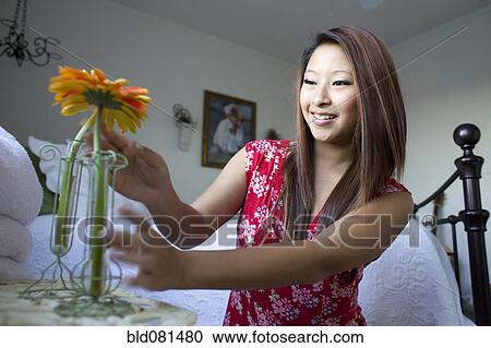 Stock Image - Smiling mixed race teenage girl placing flowers in vases on bedside table. Fotosearch
