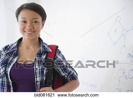 Stock Image - Smiling mixed race teenage girl standing next to whiteboard in science class. Fotosearch