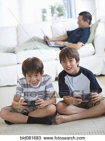 Smiling twin brothers playing video game while father uses laptop on living room sofa View Large Photo Image Stock Image - Smiling twin brothers playing video game while father uses laptop on living room sofa. Fotosearch