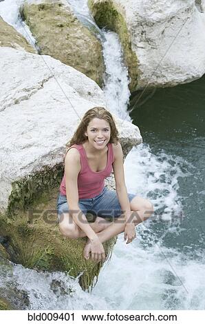 Stock Image - Woman sitting on a rock next to water. Fotosearch