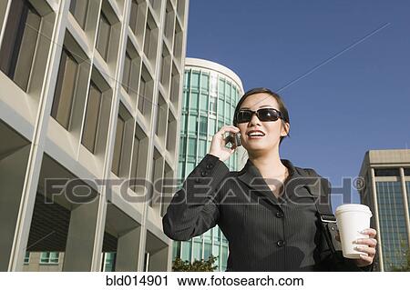 Stock Image - Businesswoman wearing sunglasses talking on cell phone with coffee. Fotosearch