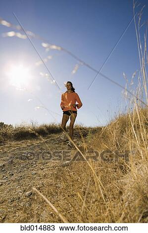 Stock Image - Full view of woman jogging on dirt road. Fotosearch