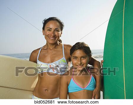 Stock Image - Hispanic mother and daughter holding surfboards. Fotosearch