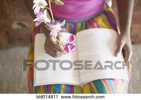 Stock Image - Low section of woman sitting in chair reading book while holding flowers. Fotosearch