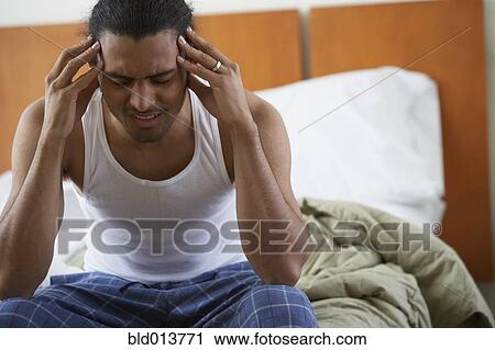 Stock Image - Man sitting on bed with hand on temples. Fotosearch
