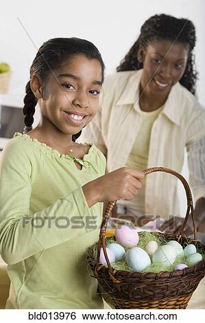 Stock Photograph - Portrait of girl holding Easter eggs in basket with mother looking at her. Fotosearch