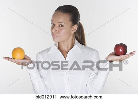 Stock Image - Portrait of woman balancing orange and apple in hands. Fotosearch