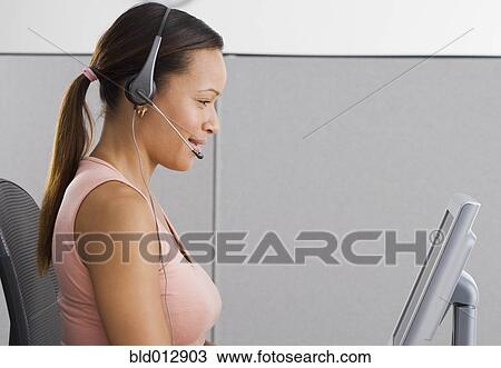 Stock Image - Side view of woman in office with headpiece in front of computer. Fotosearch
