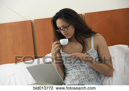 Stock Image - Woman in bed drinking coffee and looking at laptop monitor. Fotosearch