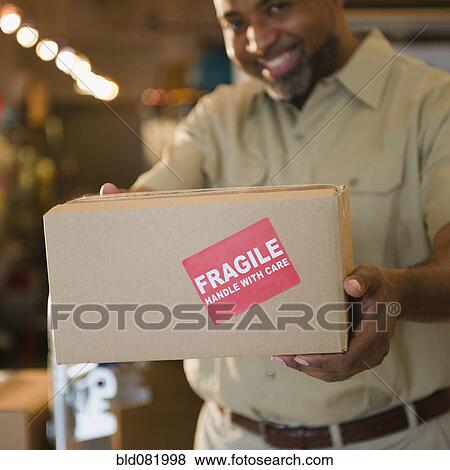 Stock Photo - African American deliveryman holding out box with 'fragile' sticker. Fotosearch