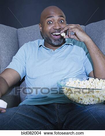 Picture - African American man eating popcorn and watching television. Fotosearch